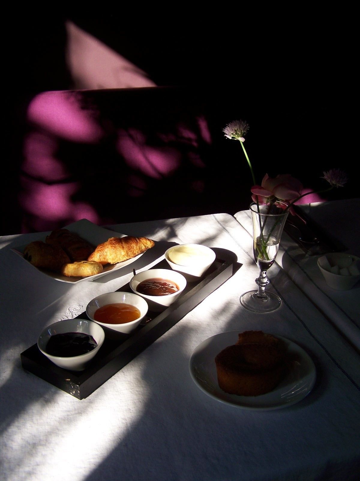 A table set with pastries, condiments, and a glass of water, with sunlight casting shadows.