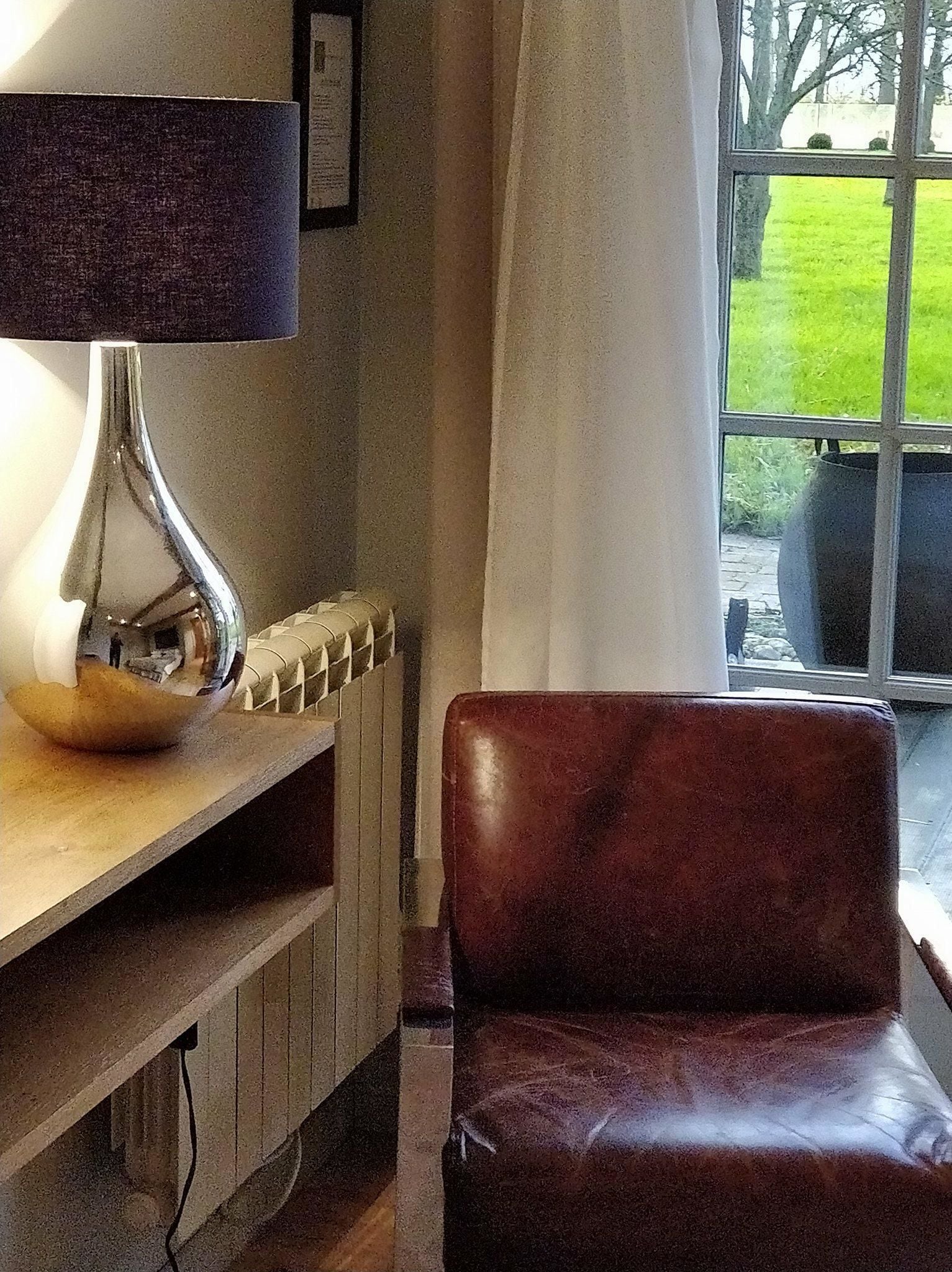 Ett rum med ett träbord, en lampa, en fåtölj i läder och en utsikt över en trädgård genom ett fönster.