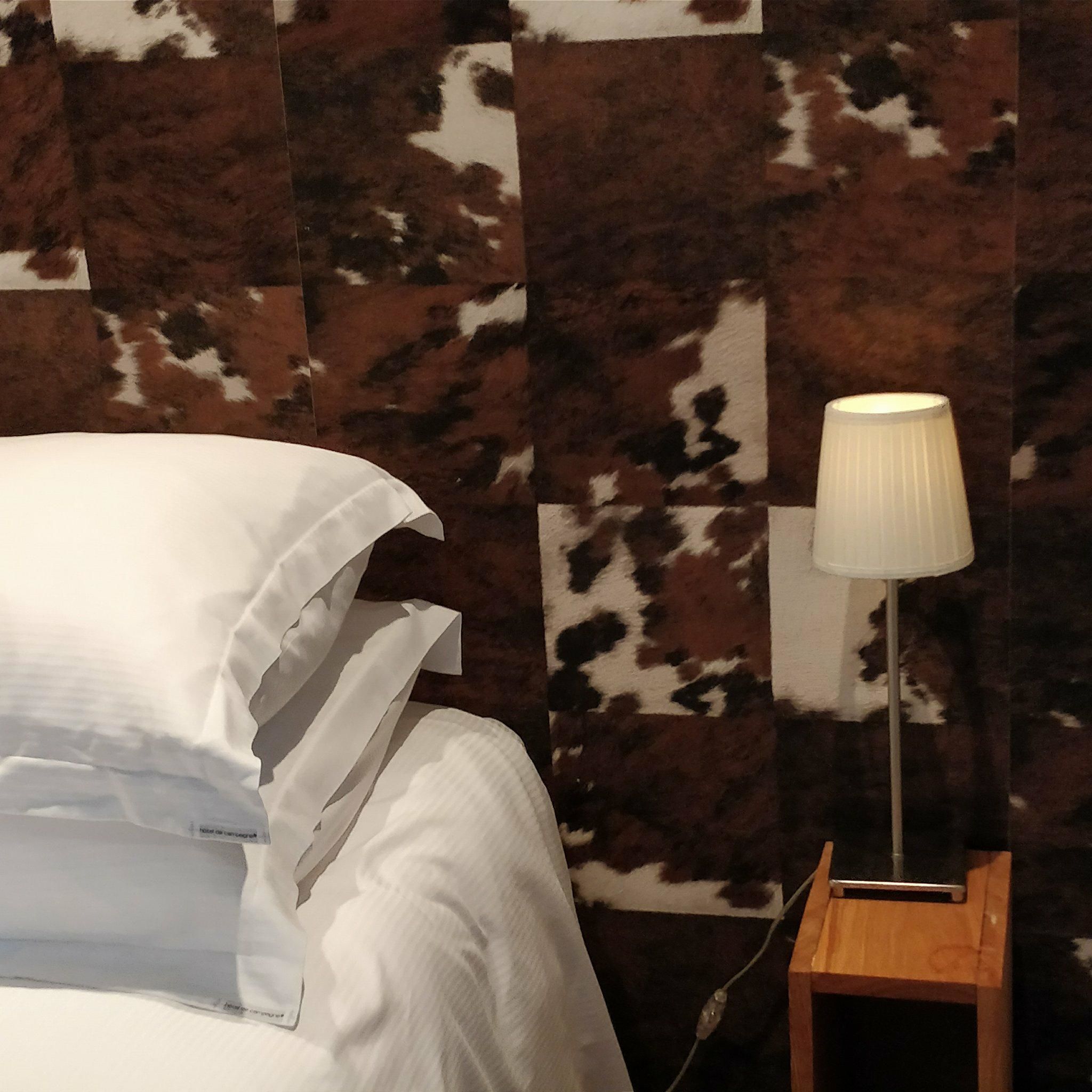 Ein Bett mit weißen Leinentüchern, ein Nachttisch aus Holz mit einer Lampe und eine Wand mit Kuhfellmuster.
