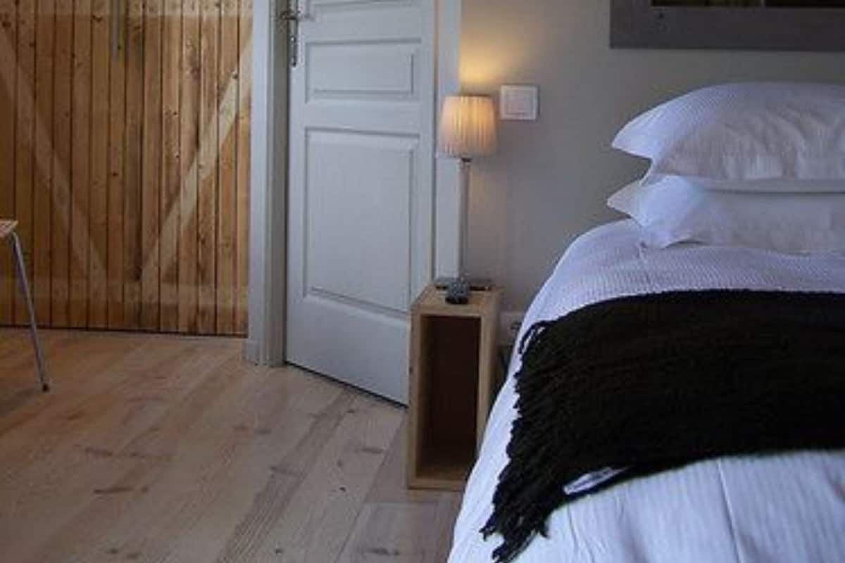 A bedroom with a wooden door, a bedside lamp, a bed with white bedding, and a window with curtains.