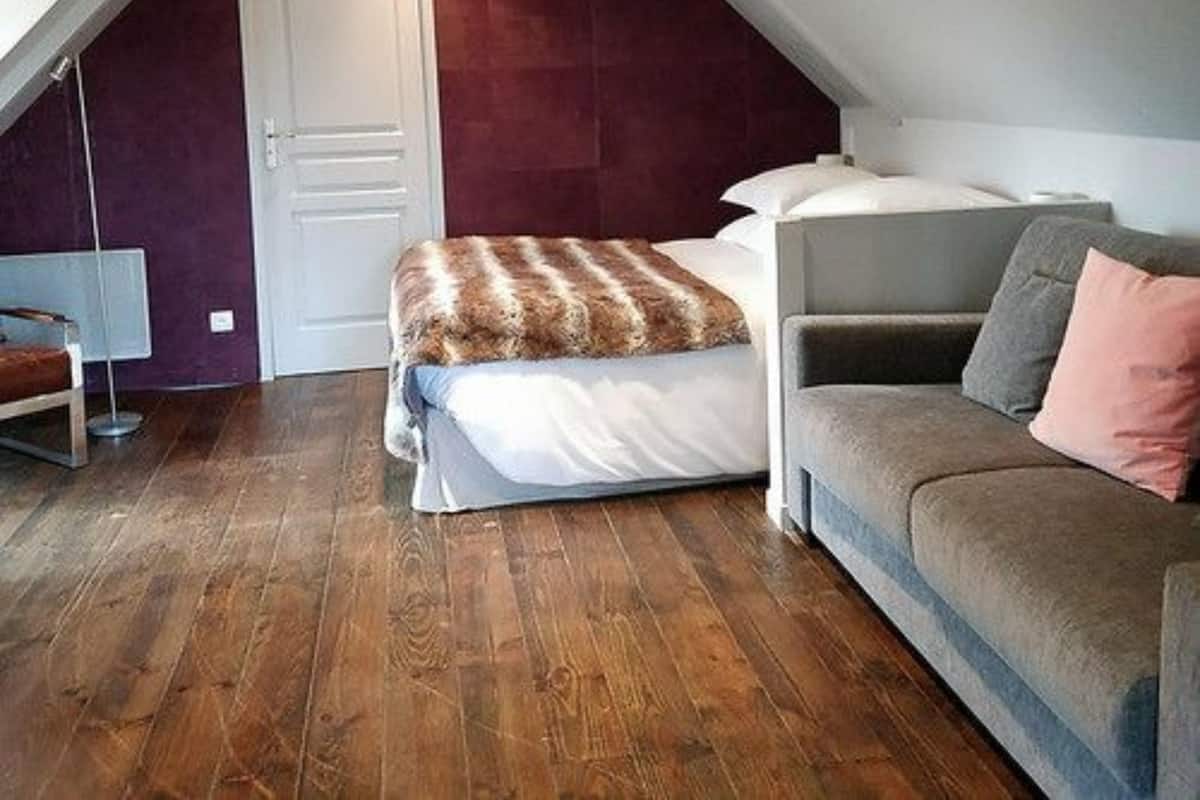 A bedroom with a sloped ceiling, a bed with a white and brown blanket, a grey sofa, and a red accent wall.