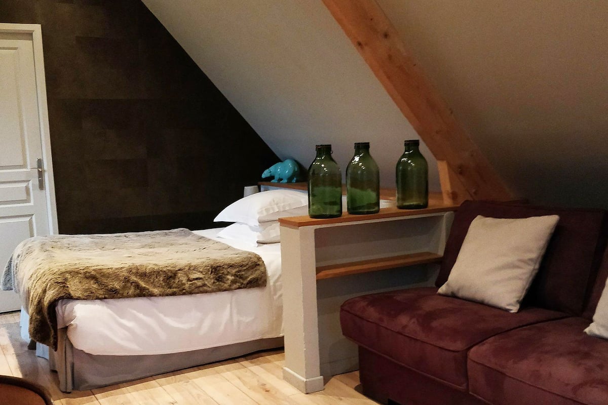 A bedroom with a bed, a wooden shelf with green bottles, a red sofa, and a white door.