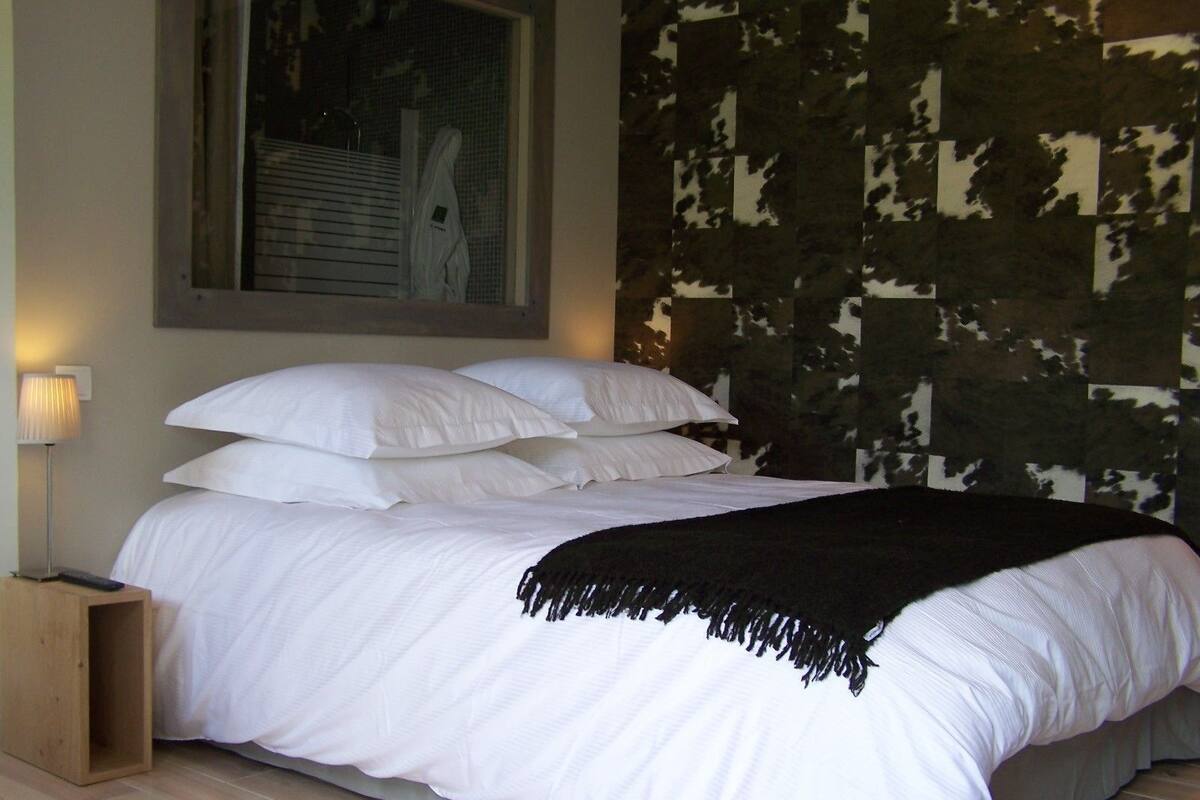 A bed with white bedding and a black throw, a bedside lamp, and a window with blinds.