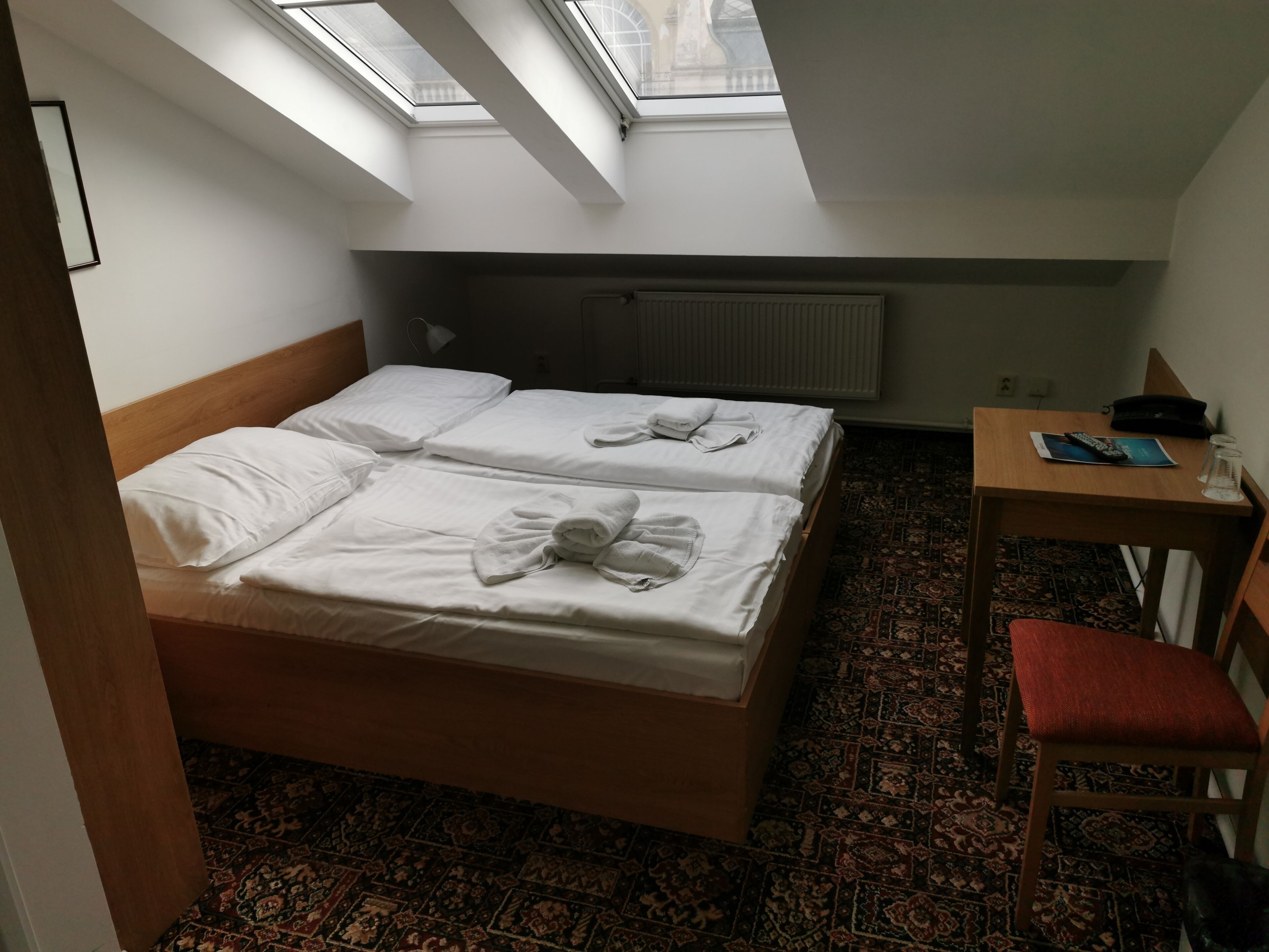A room with two beds, a small table with a chair, a radiator, and a skylight.
