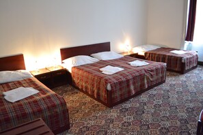 A hotel room with three single beds, each with a plaid bedspread and white pillows. There are bedside tables with lamps, and the room has patterned carpet flooring.