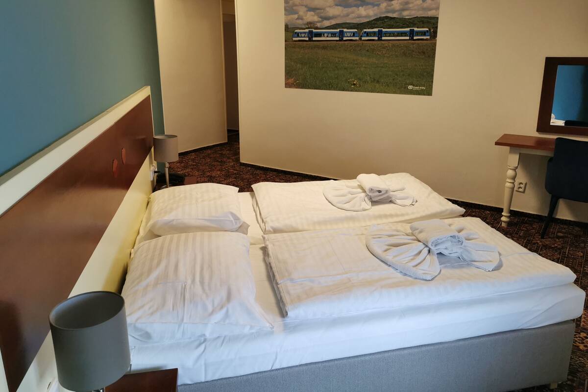 A hotel room with a bed, a desk, a chair, a lamp, and a painting of a train on the wall.