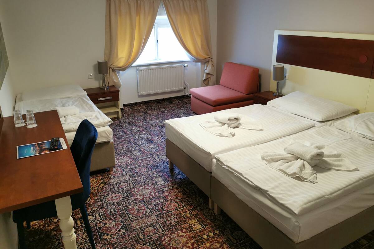 A hotel room with two beds, a desk, a chair, a red armchair, and a window with curtains.