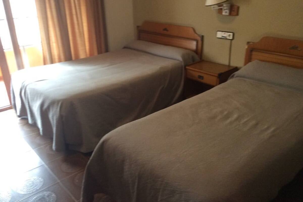 A hotel room with two single beds, a bedside table with a lamp, and a window with curtains.