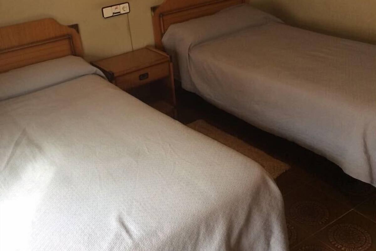 A room with two single beds, a nightstand with a lamp, and a wall-mounted control panel.