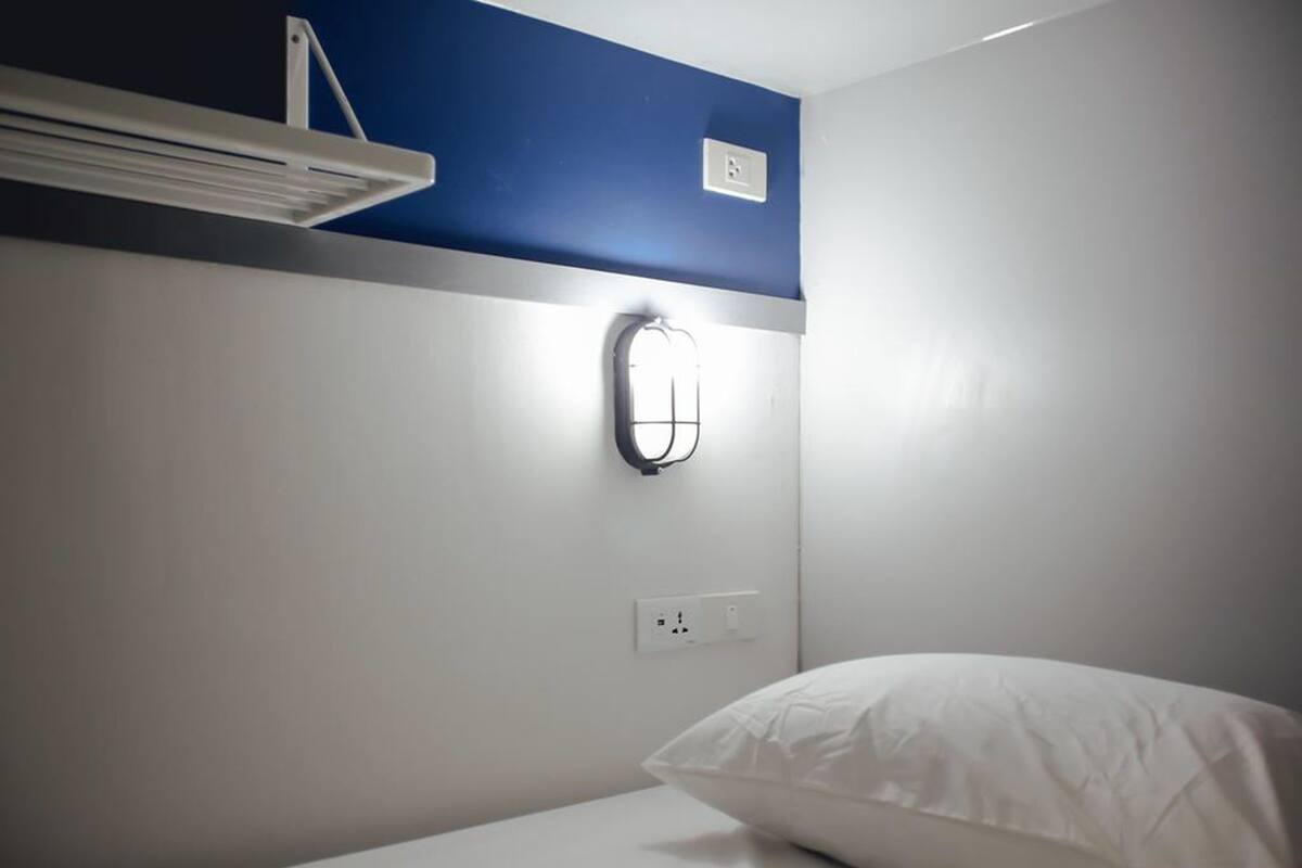 Un lit simple avec un oreiller blanc, dans une pièce dont un mur est peint en bleu, avec un luminaire fixé au mur et un ventilateur de plafond blanc.