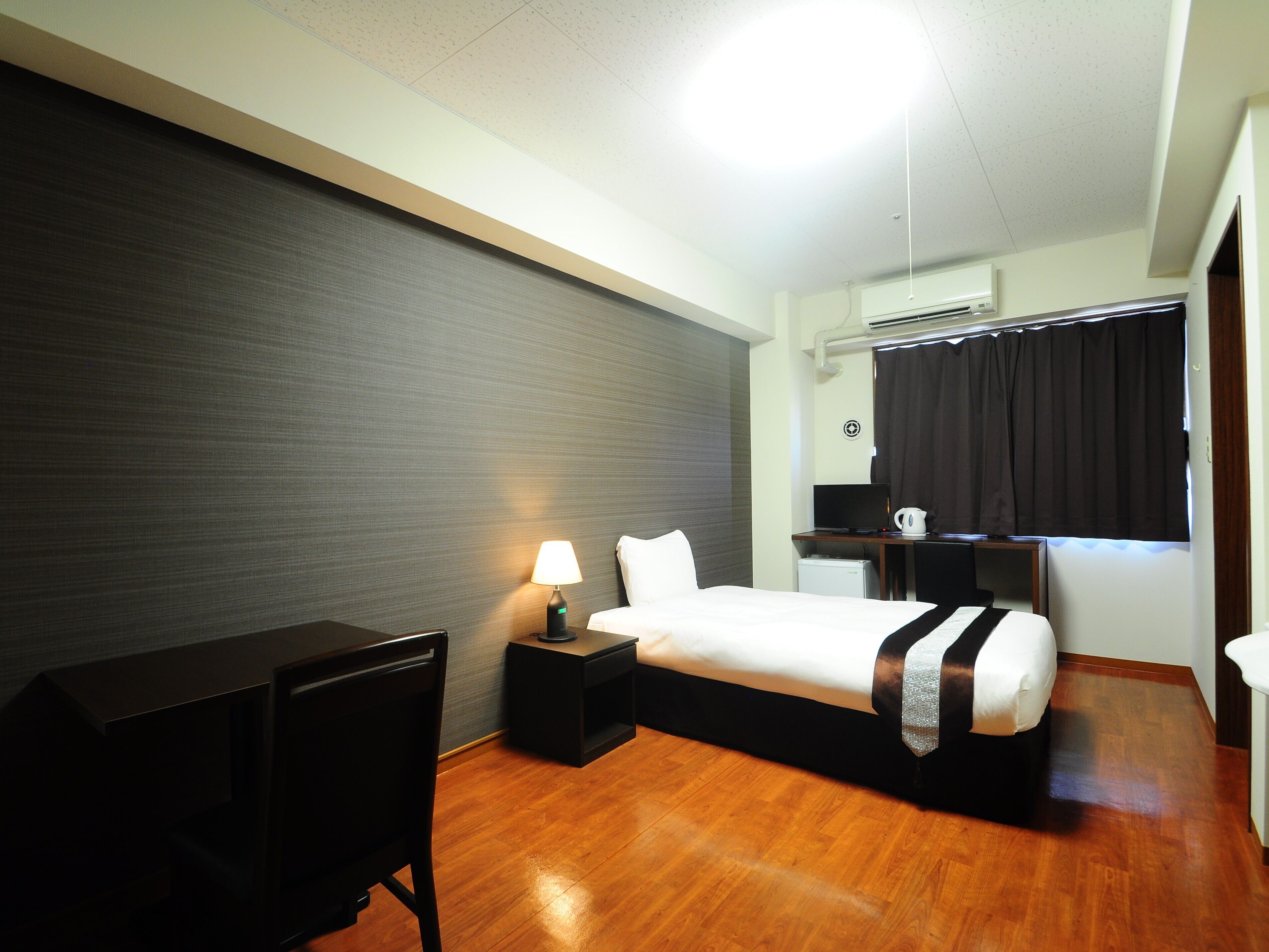 A hotel room with a bed, desk, chair, lamp, and a small kitchenette area.