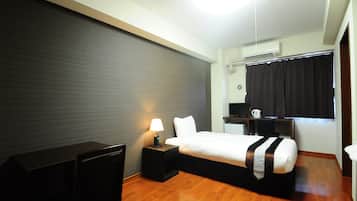 A hotel room with a bed, desk, chair, lamp, and a small kitchenette area.