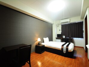 Een hotelkamer met een bed, bureau, stoel, lamp en een klein keukenhoekje.