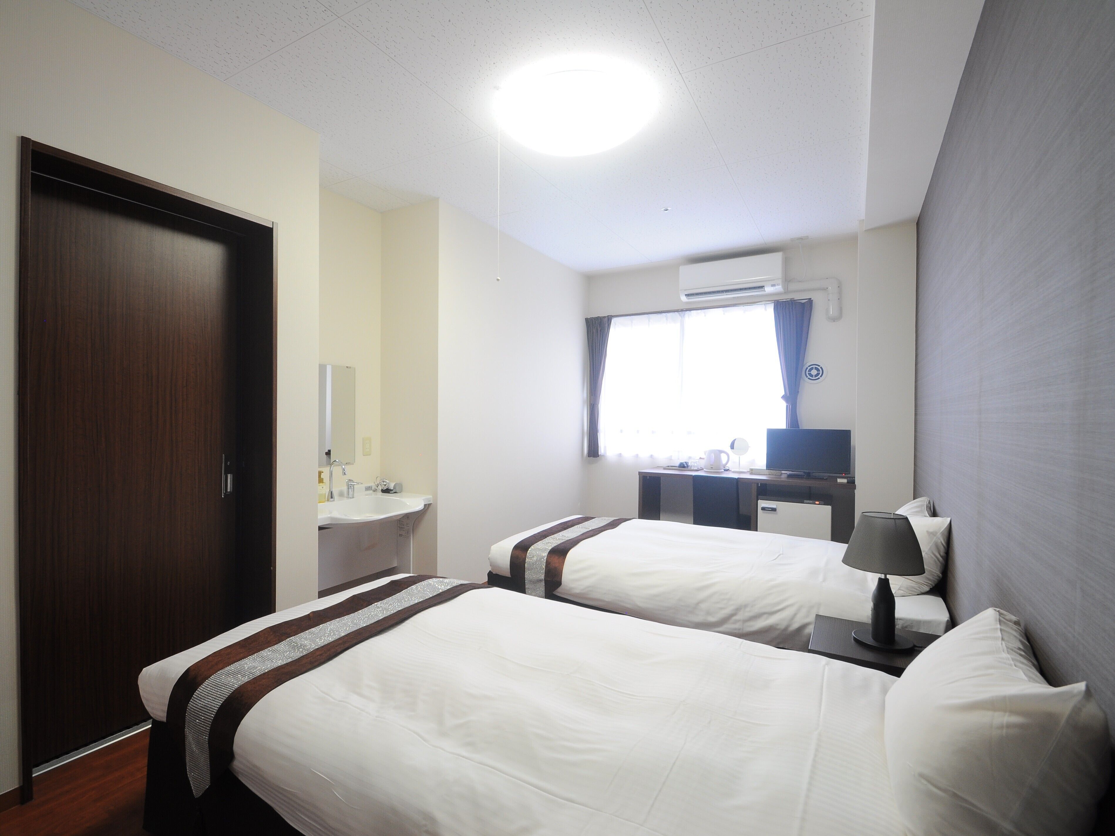 A hotel room with two beds, a desk with a flat-screen TV, a window with curtains, and an air conditioning unit.