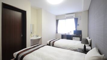 A hotel room with two beds, a desk with a flat-screen TV, a window with curtains, and an air conditioning unit.