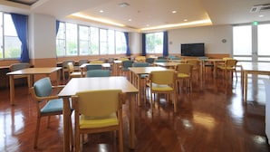 A spacious room with multiple tables and chairs, large windows, and a television on the wall.