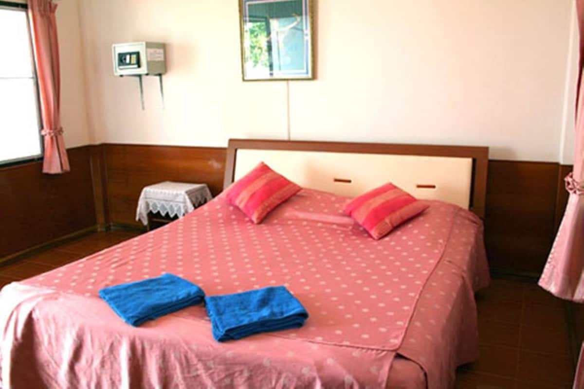 Ein ordentlich bezogenes Bett mit rosa Bettwäsche und zwei blauen Handtüchern, ein kleiner Nachttisch mit einer Lampe sowie eine wandmontierte Steckdose.