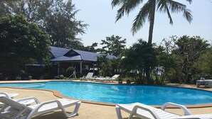A swimming pool with white lounge chairs, surrounded by trees and a building with a blue roof. - Rung Arun Resort (Ko Pha-ngan)