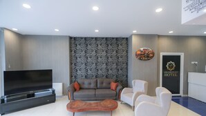 A modern hotel lobby with a grey sofa, two armchairs, a wooden coffee table, a flat-screen TV, and a reception desk. - Grand Dost Hotel (Osmancik)