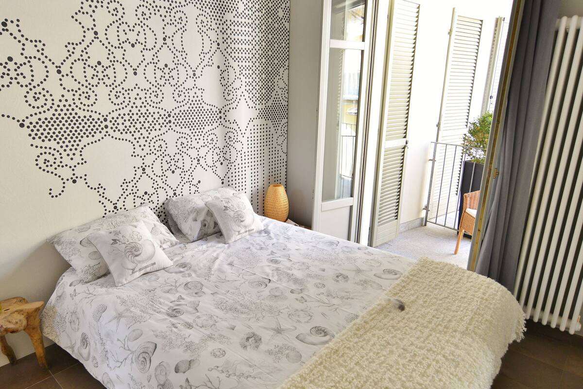 A bedroom with a bed, a nightstand, a decorative wall, and a view of the outside.
