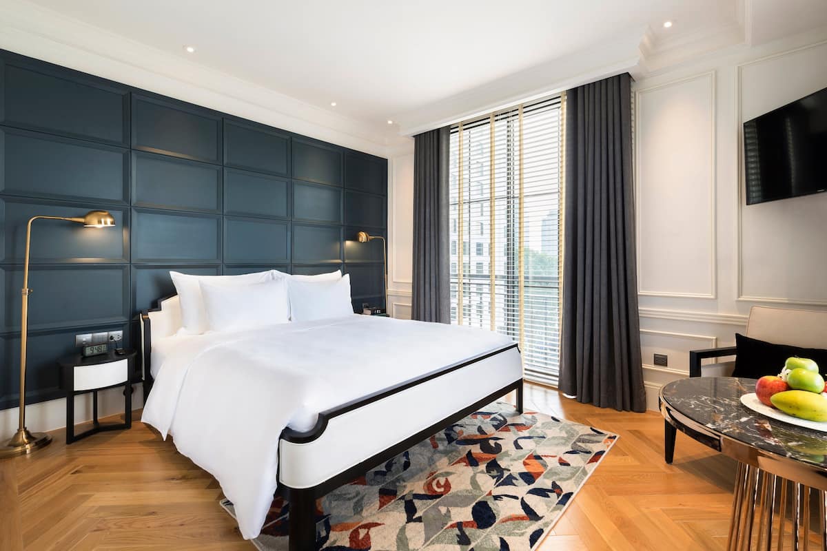 A modern hotel room with a large bed, a bedside table with a lamp, a small table with fruit, and a TV on the wall.