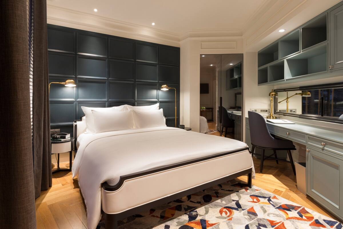 A modern bedroom with a large bed, a desk, and a chair. There is a wardrobe with a mirror and a small table with a lamp.