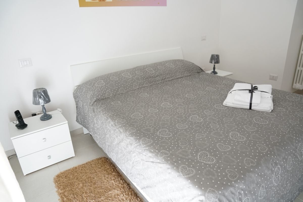 Un dormitorio ordenado con cama, mesita de noche, lámpara y una toalla doblada sobre la cama.