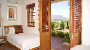 A bedroom with a bed, a nightstand, a window with blinds, and a view of a vineyard. - Lovane Boutique Wine Estate & Guesthouse (Stellenbosch)