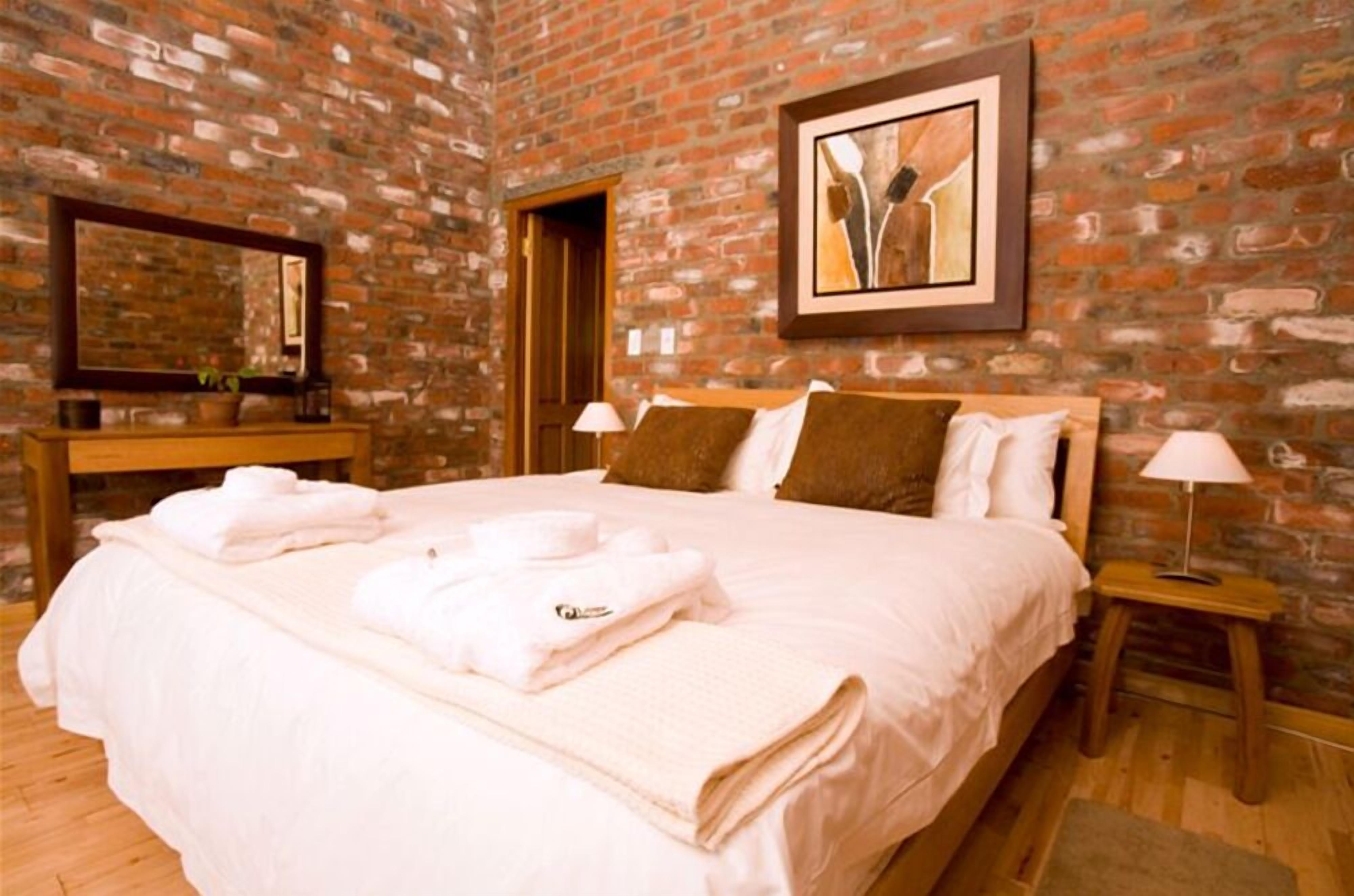 A hotel room with a brick wall, a bed with white linens, two bedside tables with lamps, and a framed abstract painting above the bed.