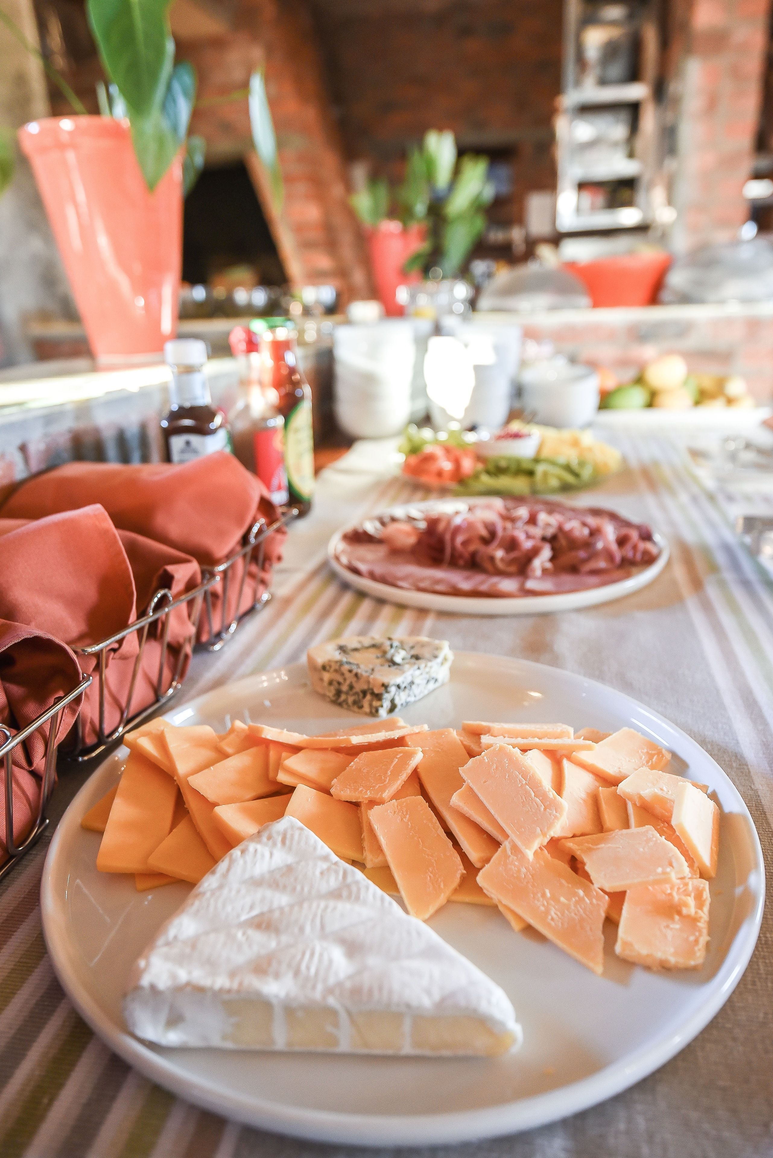A table with various cheeses and meats, a basket of napkins, and a brick wall in the background.