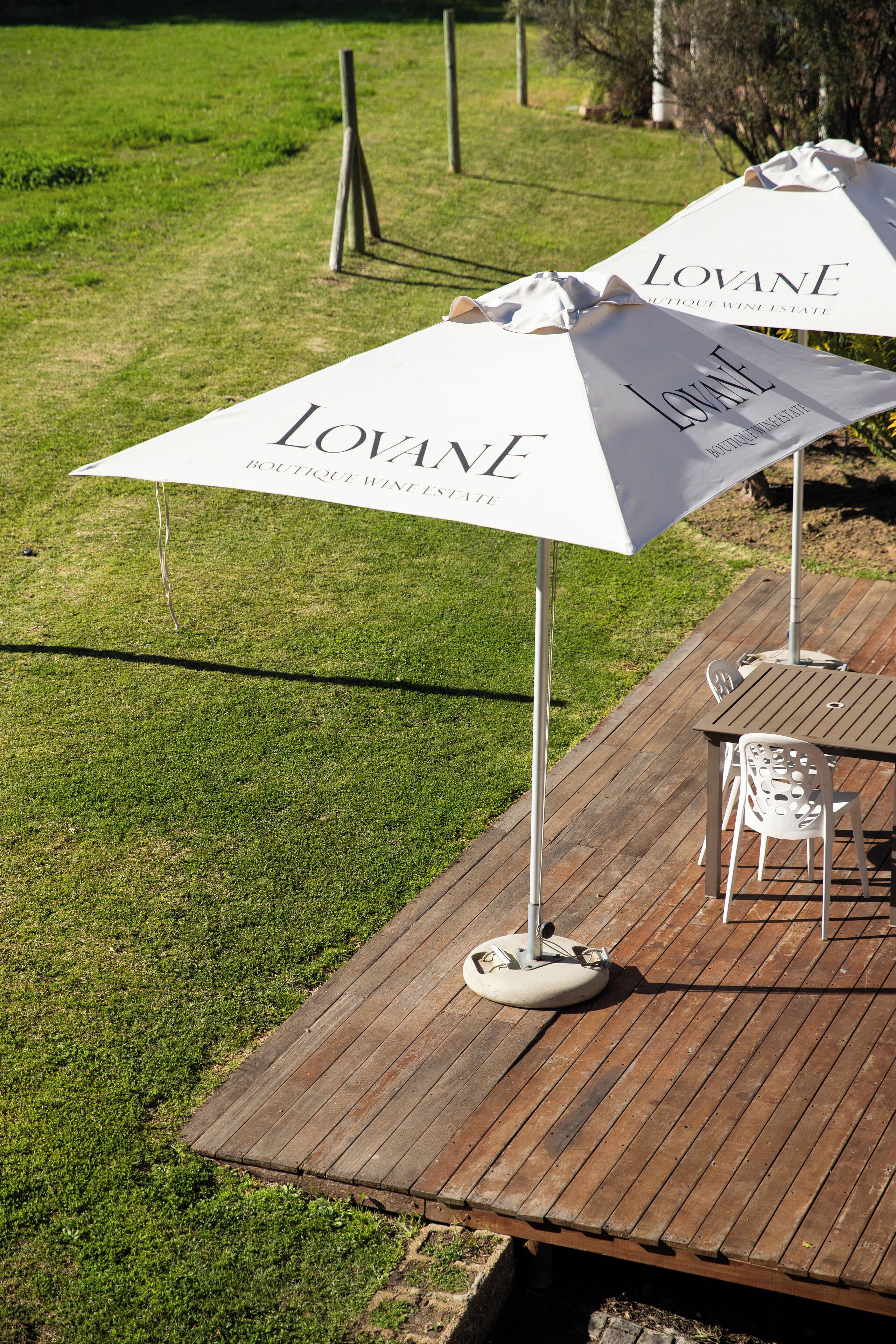 A wooden deck with two white umbrellas labeled "LOVANE BOUTIQUE WINE ESTATE" and two white chairs.