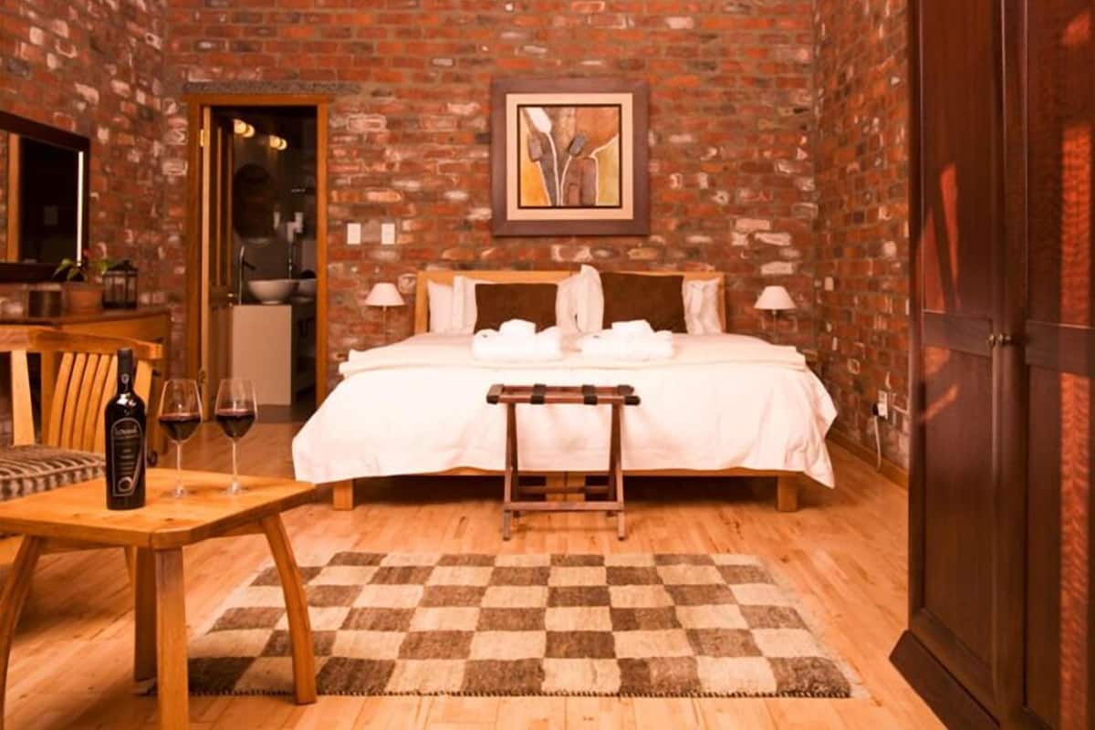 Ein Hotelzimmer mit Backsteinwand, einem Bett mit weißen Laken, einem Holztisch mit Flasche und Gläsern sowie einem Schachbrettteppich.