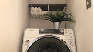 A front-loading washing machine with a digital display and control panel, located in a laundry room with a shelf above holding rolled towels and a potted plant. - Osaka guesthouse RELASTAY (Osaka)