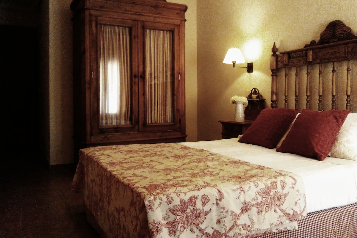 Una habitación de hotel con un armario de madera grande, una mesita de noche con lámpara y una cama con colcha estampada y dos almohadas.