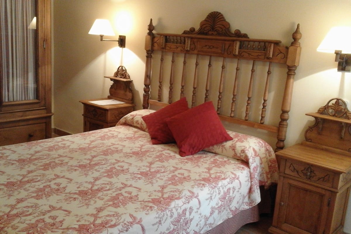 Un dormitorio con cabecera de madera, edredón estampado con flores, dos mesitas de noche con lámparas y una mesita auxiliar.