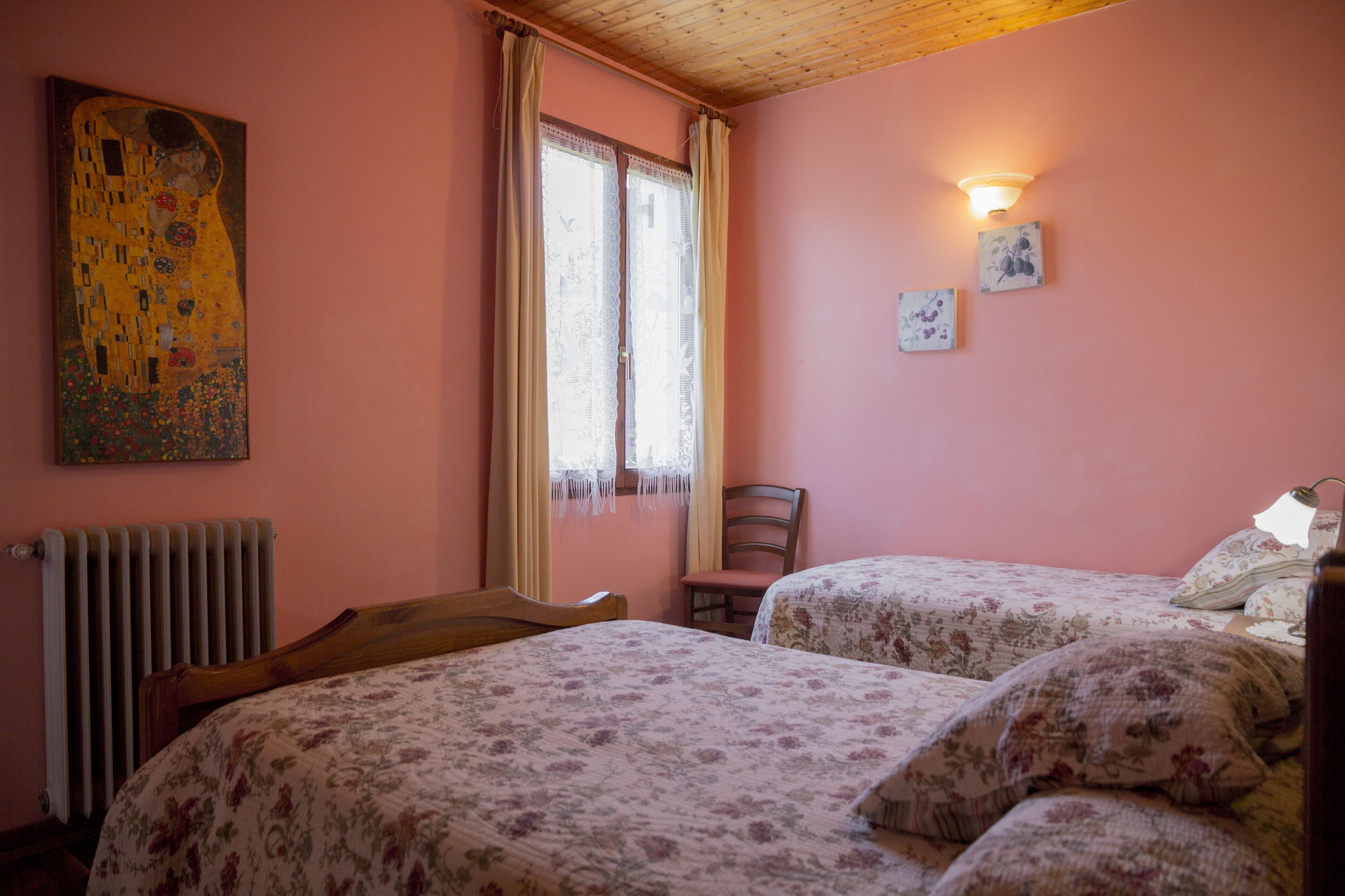 A room with two beds, a wooden headboard, a radiator, a chair, a window with curtains, and a painting on the wall.