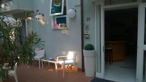 Un petit espace extérieur aménagé avec une table et des chaises, un pot de fleurs et l’entrée d’un immeuble portant le numéro 21.