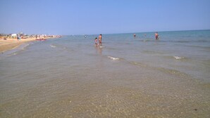 A beach with clear shallow waters, people in the water, and a distant shoreline with buildings. - Hotel Maria Gabriella (Rimini)