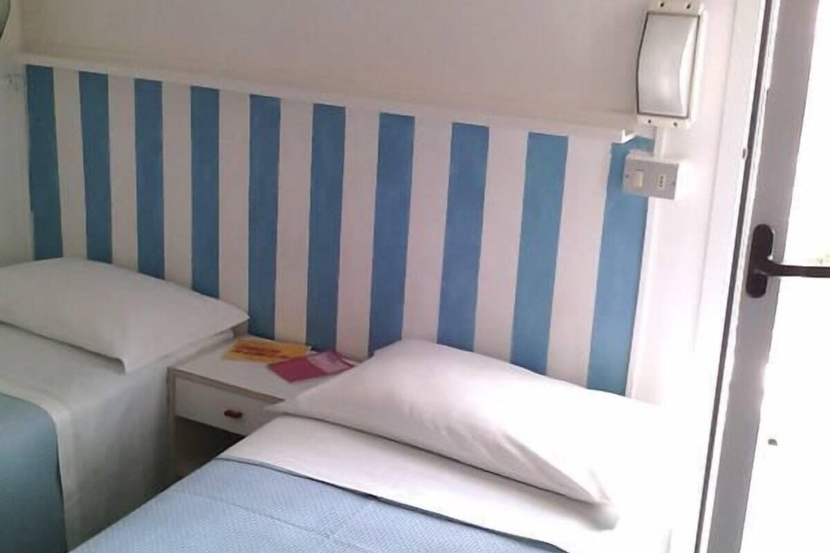 A room with two beds, each with a blue-striped headboard, a bedside table with a book, and a wall-mounted light.
