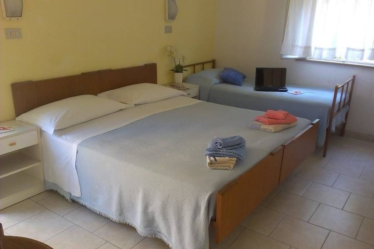 A bedroom with two beds, a bedside table, a chair, a window, and a wall-mounted lamp.