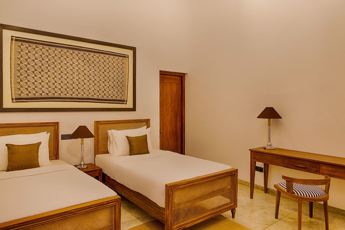 Habitación de hotel con dos camas individuales, un escritorio de madera, una silla, una lámpara y un cuadro en la pared.