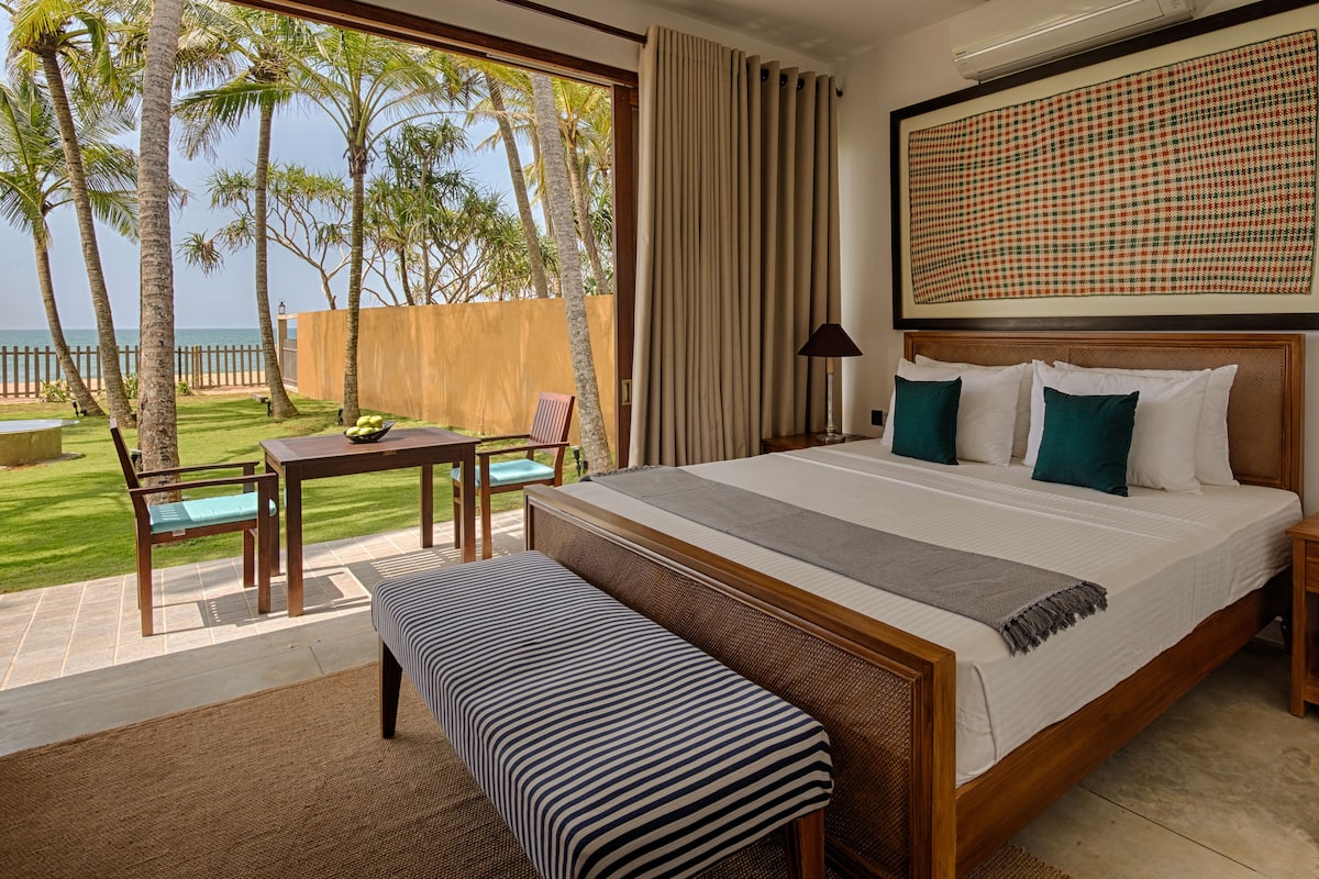 Habitación de hotel con una cama grande, una mesita de noche, un banco y vistas a palmeras y una playa.