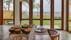 A dining table set with multiple dishes, glasses, and cutlery, overlooking a beach with a fence and palm trees. - Jetwing Villa 700 (Induruwa)