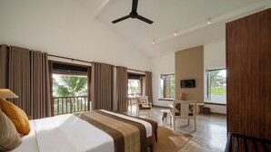 A hotel room with a bed, curtains, a desk, a chair, a television, and a balcony with a view of palm trees. - Jetwing Villa 700 (Induruwa)