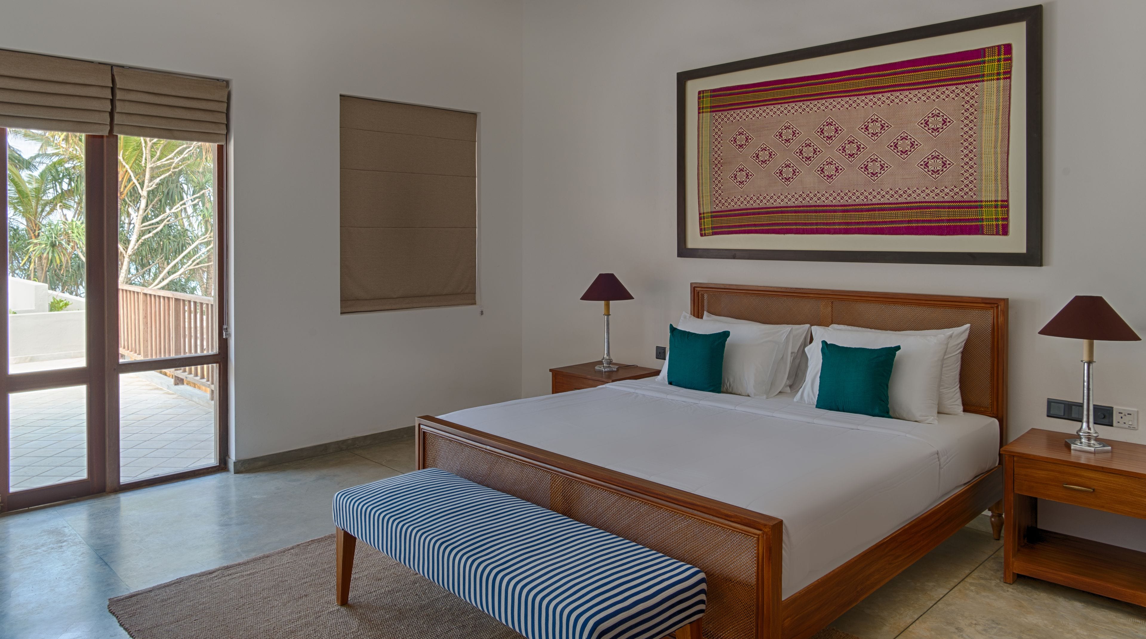 A bedroom with a large bed, two bedside tables, a bench, a window with blinds, and a wall-mounted artwork.