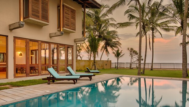 A pool with lounge chairs, palm trees, and a house with large windows. - Jetwing Villa 700 (Induruwa)