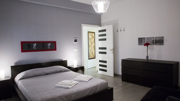 Une chambre à coucher moderne, dotée d’un grand lit, de tables de chevet, d’une commode et d’une porte donnant sur une autre pièce.