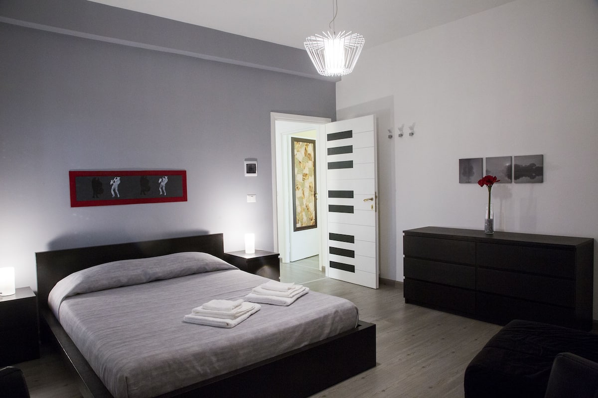 Een moderne slaapkamer met een groot bed, nachtkastjes, een dressoir en een deur naar een andere kamer.