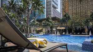 Zona de piscina con sillones de descanso, palmeras y edificios modernos al fondo.