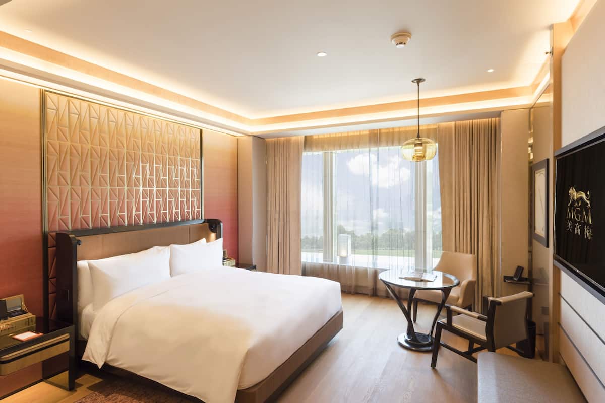 A hotel room with a large bed, a small round table, a chair, a TV, and a window with curtains.